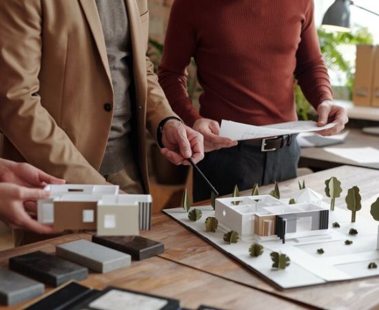 mature-male-architect-with-pencil-pointing-model-new-house-while-his-young-colleagues-holding-paper-with-sketch-part-layout_274679-18750
